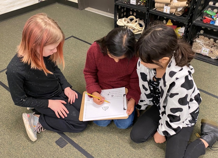 Three children gathered around a clipboard while one draws